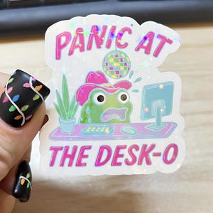 May include: A holographic sticker with the text "PANIC AT THE DESK-O". The sticker features a cartoon frog wearing a cowboy hat, looking stressed at a computer. A disco ball and a potted plant are also depicted.