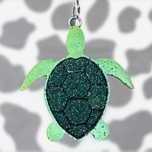 May include: A sea turtle keychain featuring a light green head and flippers with a textured surface. The central shell is a dark, glittery forest green with a distinct hexagonal pattern. A silver metal jump ring is attached to the top of the head.