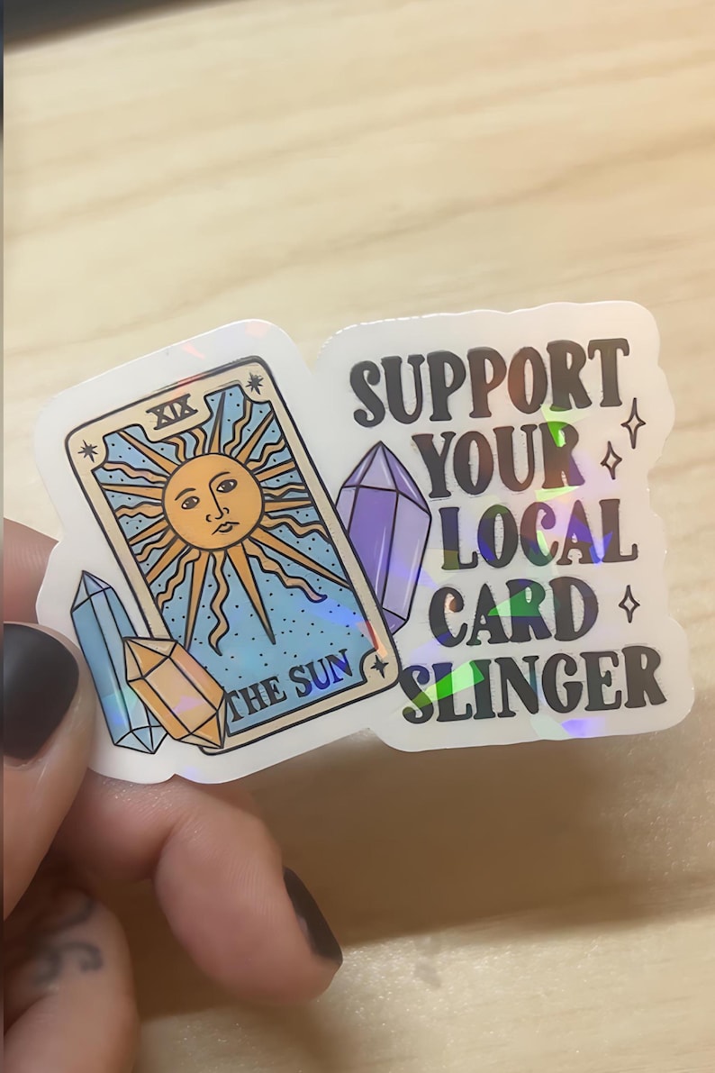 May include: A sticker featuring a tarot card design with a sun illustration and the text "THE SUN". The sticker also includes the text "SUPPORT YOUR LOCAL CARD SLINGER" and crystal graphics.