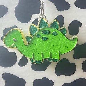 May include: A green dinosaur-shaped keychain with a gold outline and glitter accents. The keychain is attached to a silver clasp and a black and white beaded lanyard. The background is a white surface with black cow spots.