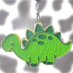 May include: A bright green dinosaur charm with a textured surface and glittery accents on the back plates and spots. The charm features a gold-toned metal outline and is attached to a silver-colored lobster clasp for easy attachment to items.