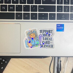 K&ouml;nnte beinhalten: Eine Laptop-Tastatur mit Aufklebern. Ein Aufkleber zeigt ein Tarotkarten-Design mit einer Sonne und dem Text "THE SUN". Ein weiterer Aufkleber lautet "SUPPORT YOUR LOCAL CARD SLINGER". Ein Intel Core i7-Aufkleber ist ebenfalls sichtbar.