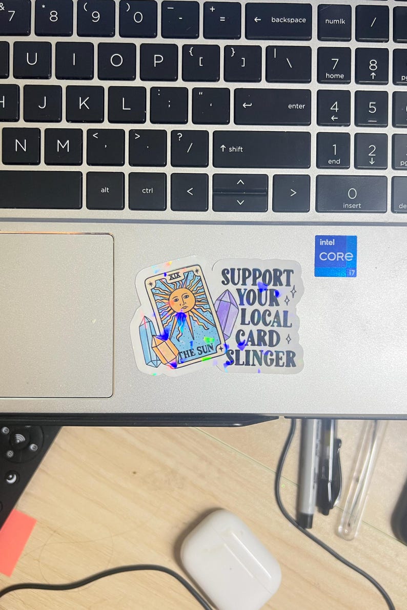 K&ouml;nnte beinhalten: Eine Laptop-Tastatur mit Aufklebern. Ein Aufkleber zeigt ein Tarotkarten-Design mit einer Sonne und dem Text "THE SUN". Ein weiterer Aufkleber lautet "SUPPORT YOUR LOCAL CARD SLINGER". Ein Intel Core i7-Aufkleber ist ebenfalls sichtbar.