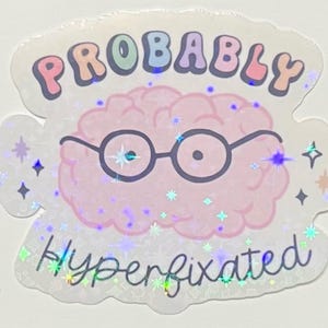 May include: A holographic sticker with a pink brain wearing glasses. The word "Probably" is arched across the top in pastel colors, and "Hyperfixated" is written below in cursive. The sticker has a sparkly, iridescent effect.