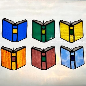 May include: A stained glass suncatcher featuring six open books arranged in two rows. The books are colored in blue, green, yellow, orange, reddish-brown, and light blue, each with a central spine detail and black leaded outlines.