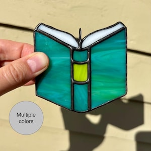 May include: A stained glass open book with teal and green hues, white page edges, and a yellow center. The book is outlined in black and has a small loop for hanging. The words "Multiple colors" are in a circle.