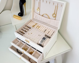 May include: A white multi-level jewelry box with a hinged lid, featuring a velvet-lined interior with compartments for rings, necklaces, and earrings. The box includes a pull-out drawer, a gold-tone latch, and a watch displayed on a cushion.