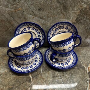 May include: Four ceramic teacups and saucers with a blue floral pattern. The cups and saucers are white with a blue floral design around the edges. The cups have a blue handle. The set is on a marble surface.
