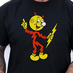 May include: Black t-shirt featuring a cartoon character with a yellow head, red body, and yellow shoes. The character holds a yellow lightning bolt and has a bubble coming from its mouth. The shirt is worn by a person holding a white mug.