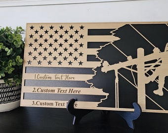 May include: Wooden American flag with a silhouette of a lineman working on a power pole. The flag is painted in shades of brown and black. The text "1. Custom Text Here", "2. Custom Text Here", and "3. Custom Text Here" is engraved on the flag.