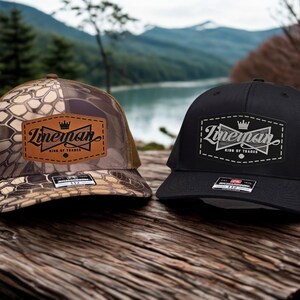 May include: Two trucker caps, one camouflage and one black, both featuring the word "Lineman" and a crown logo on a leather patch. The caps are displayed on a wooden surface, with a scenic background.