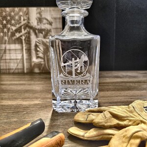 Personalized Lineman whiskey decanter engraved with the name Rivera on a rustic display with lineworker art, tools, and work gloves.