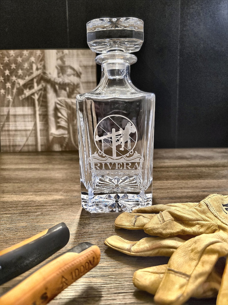 Personalized Lineman whiskey decanter engraved with the name Rivera on a rustic display with lineworker art, tools, and work gloves.
