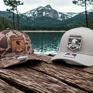 May include: Two trucker hats with the word "FREEDOM" on a patch. One hat has a camouflage pattern, the other is gray. Both have a mesh back and a patch with an eagle and the text "250 Years".