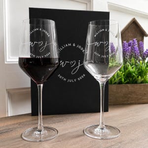 May include: Two clear glass wine glasses with long stems. One glass is filled with red wine. Both glasses feature white etched text reading William & Jodee, 30th July 2026, and a stylized W and J with a heart symbol in the center of the bowl.