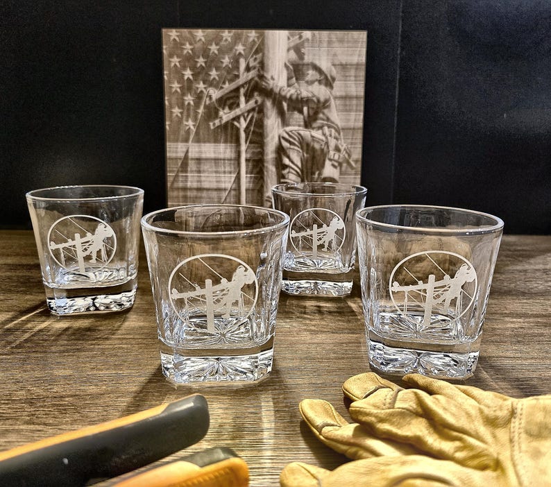 Four rocks glasses engraved with the standard Lineman emblem on a rustic display with lineworker art, tools, and work gloves.