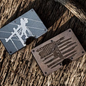 May include: Two rectangular metal wallets with rounded corners and exposed screws. One wallet is dark gray with a white graphic of a utility worker on a pole. The other wallet is brown with a wood-grain texture and a dark engraved American flag design.