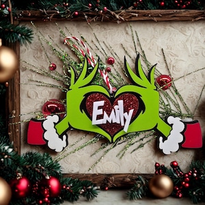 May include: A festive Christmas decoration with a Grinch-inspired hand design in green, forming a heart shape. The heart is red and features the name "Emily" in white. The decoration is surrounded by Christmas ornaments and foliage.