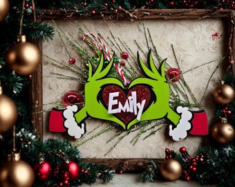 May include: A Christmas-themed decoration featuring a green Grinch-like hand design forming a heart shape. The heart is red with the name "Emily" in white. The decoration is surrounded by Christmas ornaments and greenery.