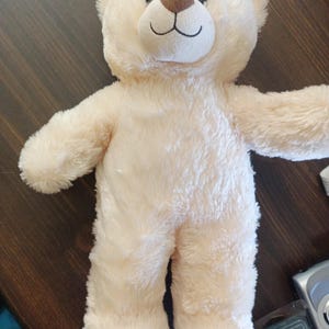 Op de afbeelding: Een crème-kleurige teddybeer met een bruine neus en een gestikte zwarte glimlach. De beer heeft zachte, donzige vacht en uitgestrekte armen, wat wijst op een knuffelbaar ontwerp. Een klassiek kinderspeelgoed.