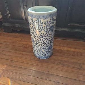May include: A tall, cylindrical ceramic umbrella stand. The stand is white with a blue floral and abstract pattern. The rim has a blue heart design. The interior is a light blue color.