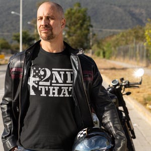 May include: Black t-shirt with the text "2ND THAT" in white, featuring a rifle design with stars. The person is wearing a black leather jacket and holding a motorcycle helmet.