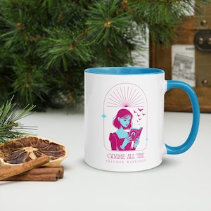 May include: A white ceramic mug with a light blue rim and handle. The mug features a pink and blue illustration of a person reading a book with bats flying around them. The text "Gimme all the trigger warnings" is printed in pink at the bottom of the mug.