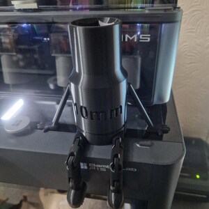 May include: A black 3D printed figurine of a robot with articulated arms and legs. The robot is sitting on a black surface and has a 10mm diameter cylinder for a body. The robot is labelled "10mm" on the cylinder.