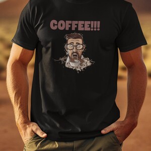 Puede incluir: Una camiseta negra con una ilustración de dibujos animados de un hombre con una expresión de sorpresa, usando gafas y bebiendo café. El texto "COFFEE!!!" está impreso en marrón encima de la ilustración.