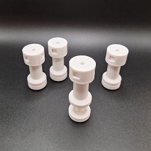 May include: Four white, cylindrical objects, each resembling a dumbbell, are displayed on a dark surface. They have wider top and bottom sections joined by a narrower central part. The top section of each has a square indentation.
