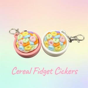 May include: Two cereal-themed fidget toys. One is in a pink bowl, the other in a white bowl. Both are filled with colourful, ring-shaped cereal pieces. The text "Cereal Fidget Cickers" is written in pink cursive.
