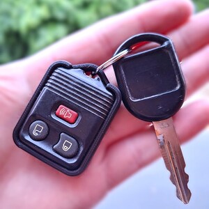 May include: A black automotive key fob with three buttons, including one red panic button and two black lock and unlock buttons, attached to a metal key ring with a standard black plastic-headed ignition key featuring a metallic blade.
