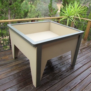 Puede incluir: Un arriate elevado hecho de madera con un acabado beige claro y un borde gris oscuro. El arriate está sentado en una plataforma de madera.