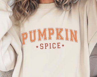 May include: Cream-colored sweatshirt with the text "PUMPKIN SPICE" in orange block letters. The word "SPICE" is below "PUMPKIN" and is separated by a star. The sweatshirt is a casual style, perfect for fall.