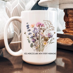 May include: White ceramic mug with a floral design featuring dried flowers in shades of purple, pink, and white. The mug has a black handle and the text "His mercies are new every morning" printed on the bottom.
