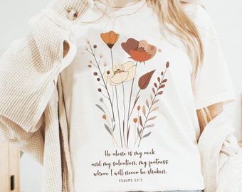 May include: White t-shirt with a floral design and the text "He alone is my rock and my salvation, my fortress where I will never be shaken. Psalms 62:7"