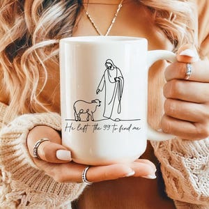 May include: White ceramic mug featuring a line drawing of a figure with a sheep, with the text "He left the 99 to find me." The mug has a large handle and is held by a person wearing a cream-colored sweater.