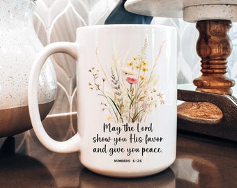 Könnte beinhalten: Eine weiße Keramiktasse mit einem Blumenmuster und dem Text "Möge der Herr dir seine Gunst erweisen und dir Frieden geben. Numeri 6:26"