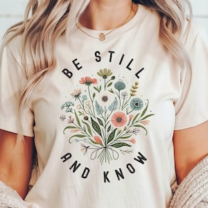 May include: A soft cream colored t-shirt with a floral design and the text "Be Still and Know" printed on the front.