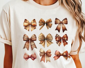 Puede incluir: Camiseta color crema con nueve lazos decorativos de varios patrones y colores. Los lazos incluyen diseños de cuadros, lunares y lisos en tonos marrón, dorado y rojo. La camiseta tiene mangas cortas y un corte holgado.