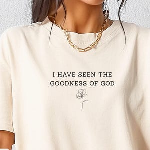 May include: Ivory-colored t-shirt with the text "I HAVE SEEN THE GOODNESS OF GOD" in black, along with a minimalist floral illustration. The shirt has a classic crew neck and short sleeves, suitable for casual wear.