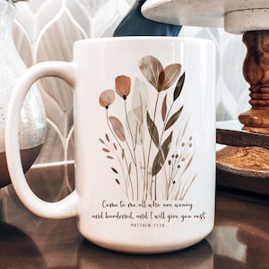 May include: White ceramic mug with a floral design in shades of brown, green, and pink. The mug has a quote from Matthew 11:28 that reads "Come to me all who are weary and burdened, and I will give you rest."