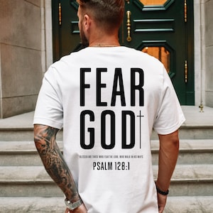 Op de afbeelding: Wit T-shirt met de tekst "FEAR GOD" in grote, vette, zwarte letters op de achterkant, samen met een kruis en Psalm 128:1. Het shirt heeft een klassieke ronde hals.