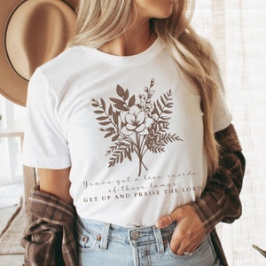 May include: White t-shirt with a floral design and the text "You've got a lion inside of these lungs GET UP AND PRAISE THE LORD".