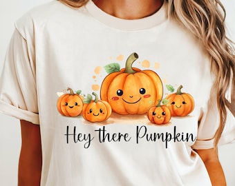 Puede incluir: Camiseta beige con una ilustración de acuarela de cinco calabazas sonrientes. Las calabazas son naranjas con tallos y hojas verdes. El texto "Hey there Pumpkin" está escrito en escritura negra debajo. Diseño informal con temática otoñal.