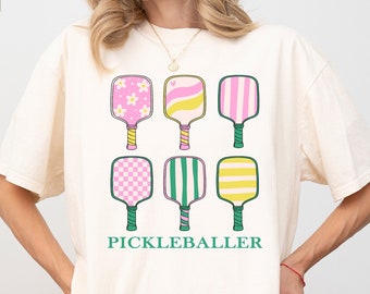 Könnte beinhalten: Ein weißes T-Shirt mit einem Aufdruck von sechs verschiedenen gemusterten Pickleball-Schlägern und dem Text "Pickleballer" in Grün.