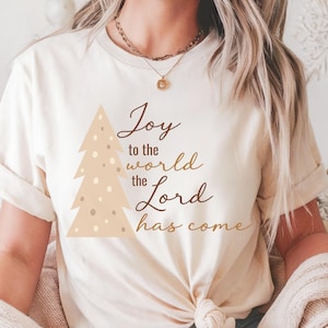May include: A cream-coloured t-shirt with a light brown Christmas tree design and the text "Joy to the world the Lord has come". The festive tee features a script font and is suitable for the holiday season.