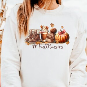 May include: White sweatshirt with a fall-themed graphic. The design includes brown boots, a coffee cup, a knit hat, and a pumpkin with a pink bow. The text "#FallBasics" is also included in the design.