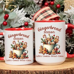May include: Two white ceramic mugs with red interiors and handles. The mugs feature a "Gingerbread season" design with gingerbread cookies, candy canes, and text. The mugs are on a wooden surface with Christmas decorations.