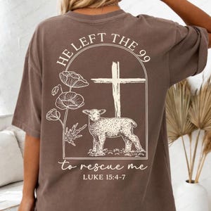 May include: A brown t-shirt with a graphic on the back. The design includes a cross, a lamb, and floral illustrations. The text reads "HE LEFT THE 99 to rescue me" and "LUKE 15:4-7". The shirt has short sleeves and a loose fit.